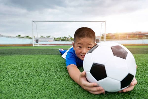 Boy lying on artificial turf with a football in front of a goal