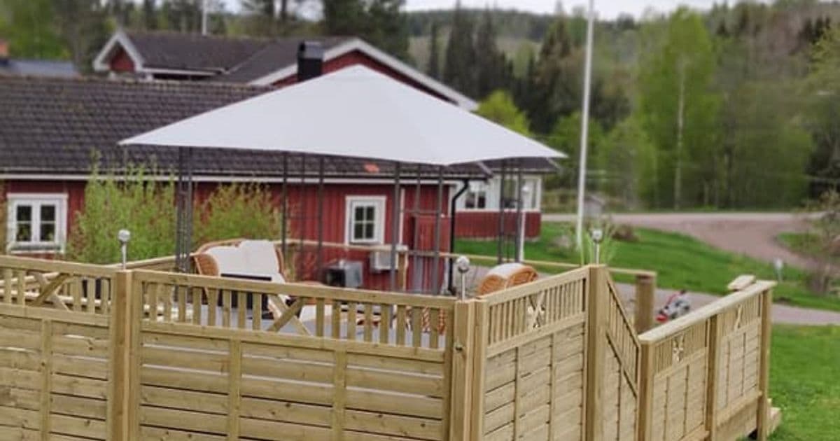 Inhägnat trädäck med paviljong och utemöbler framför ett rött hus